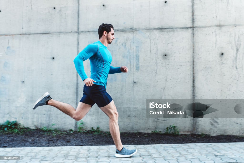 Young man running outdoors in morning Side view of young man running outdoors in morning. Male athlete in running outfit sprinting outdoors. Running Stock Photo