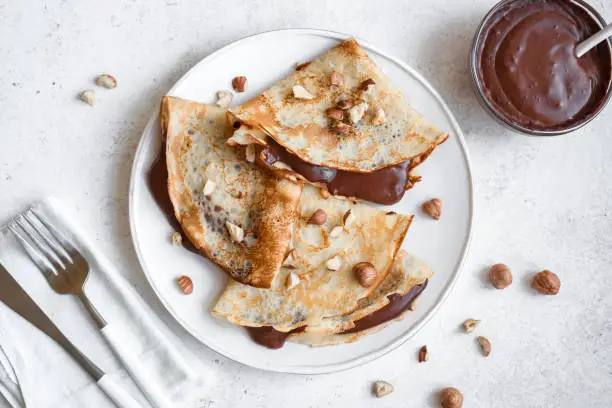 Crepes with chocolate spread and hazelnuts. Homemade thin crepes for breakfast or dessert on white background, copy space.