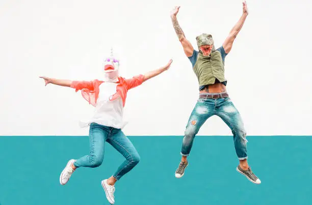Happy seniors crazy couple wearing unicorn and t-rex mask while jumping outdoor - Mature trendy people having fun celebrating outside - Absurd concept of masquerade funny holidays
