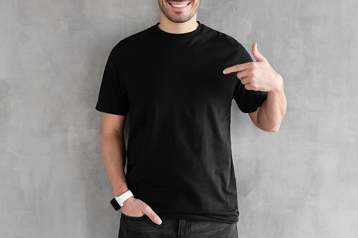 Hombre joven aislado en una pared con textura gris, sonriendo mientras apunta con el dedo índice a camiseta negra, copyspace para publicidad photo