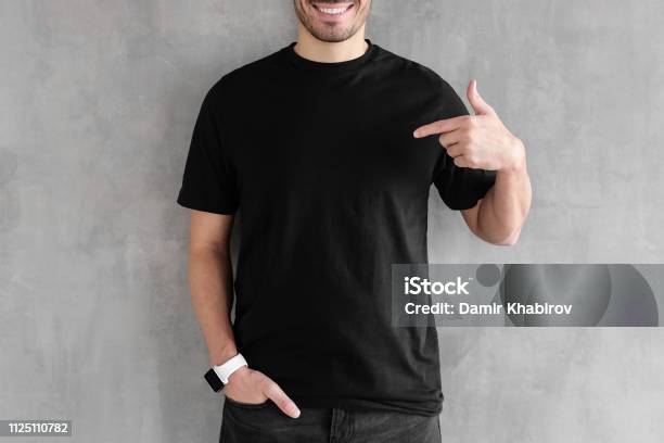 Young Man Isolated On Gray Textured Wall Smiling While Pointing With Index Finger To Black Tshirt Copyspace For Advertising Stock Photo - Download Image Now