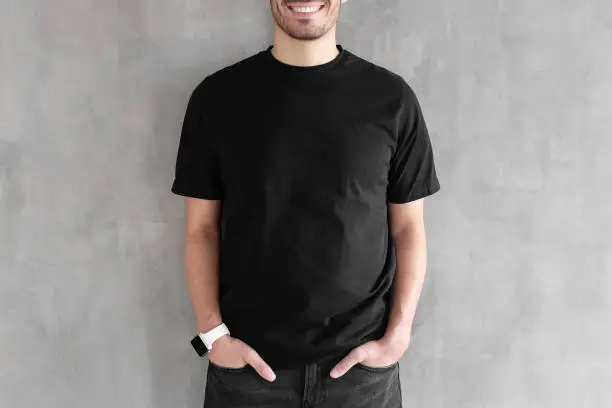 Hotizontal portrait of young man wearing blank black t-shirt and jeans, posing against gray textured wall