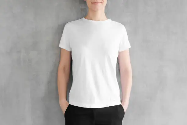 Young woman posing in blank white cotton t-shirt, standing against gray textured wall. No face photo