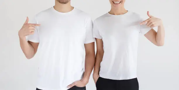 Daylight shot of smiling couple pointing at  blank white t-shirts with index finger, copy space for ads, isolated on gray background