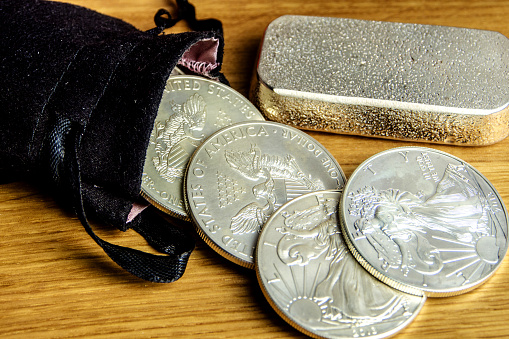 closeup of silver coins american one ounce eagles and brick falling out of black money bag and laying on wooden background