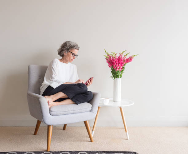 Middle aged woman with grey hair and glasses sitting  in retro armchair using smart phone