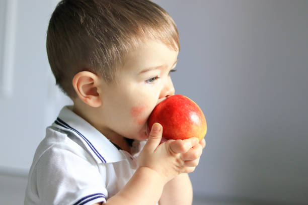 Close up portrait of cute little baby boy with atopic dermatitis on his cheek holding and eating red apple. Food allergy stock photo