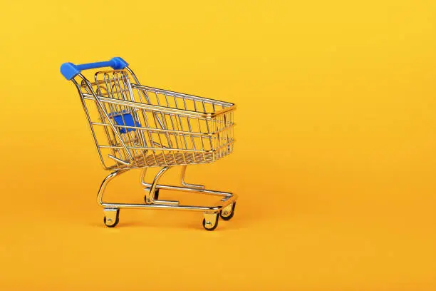 Close up empty toy metal supermarket shopping cart over vivid yellow background with copy space, low angle side view