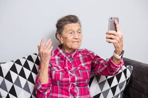 灰色の髪と深い非常に古い上級白人おばあさんしわホーム ジーンズと赤い格子縞のシャツのソファに座っていると彼女のスマート フォンを使用して、ビデオ通話用のフロント カメラを伸ば� - love life ストックフォトと画像