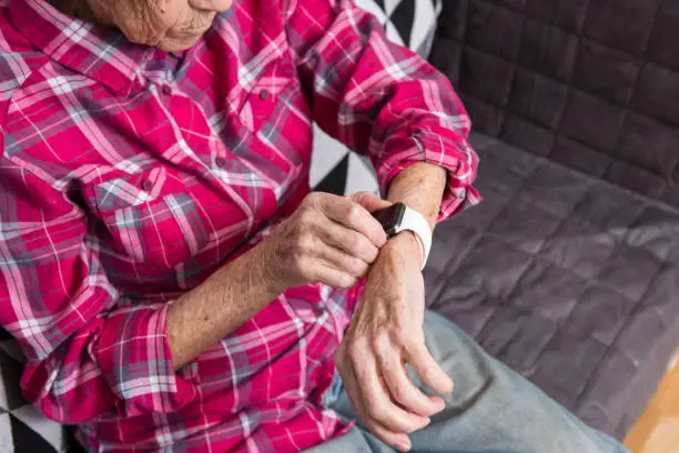 Photo of A very old senior Caucasian grandmother with gray hair and deep wrinkles sits at home on the couch in jeans and a red plaid shirt and uses a smart watch on her wrist. Pensioner and technology