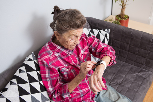 A very old senior Caucasian grandmother with gray hair and deep wrinkles sits at home on the couch in jeans and a red plaid shirt and uses a smart watch on her wrist. Pensioner and technology.