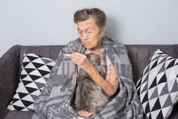 bardzo stara starsza babcia rasy kaukaskiej z siwymi włosami i głębokimi zmarszczkami siedzi w domu na kanapie z kotem domowym. choroba jest przeziębienia i gorączka. emeryt trzyma termometr w rękach - one person lifestyles 80 plus years indoors zdjęcia i obrazy z banku zdjęć