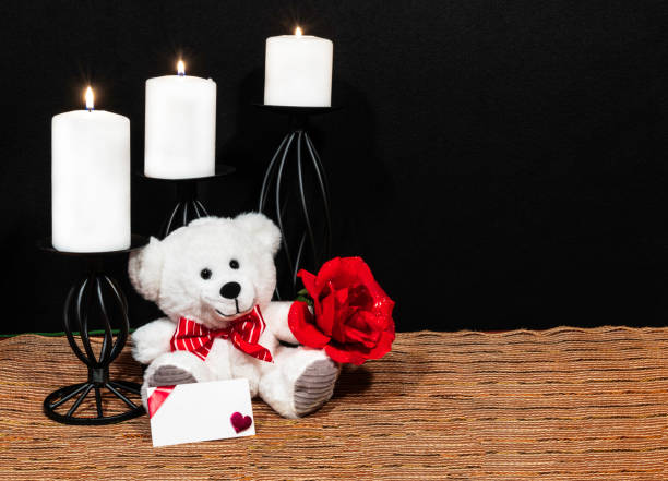 istock Cuddly teddy bear with red bow tie, red rose, white candles perched on black candle holders on mesh place mat and wooden table with card and dark background. 1125191111