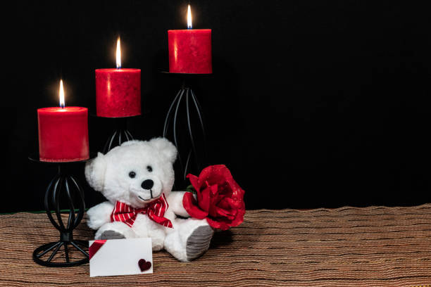Cuddly teddy bear with red bow tie, red rose, red candles perched on black candle holders on mesh place mat and wooden table with card and dark background.