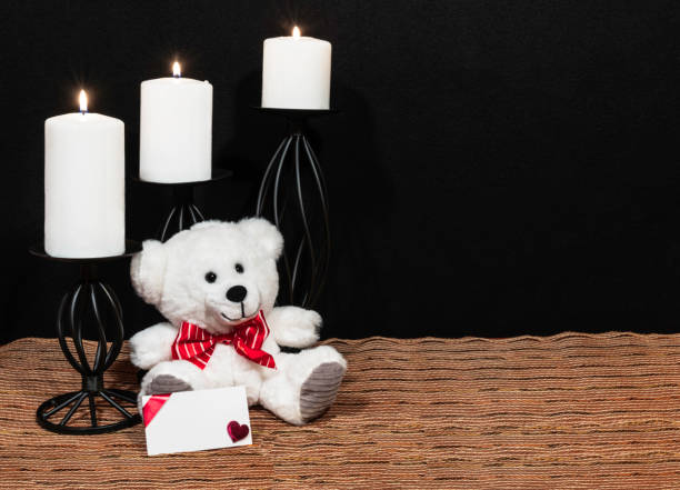 istock Cuddly teddy bear with red bow tie, white candles perched on black candle holders on mesh place mat and wooden table with card and dark background. 1125191150