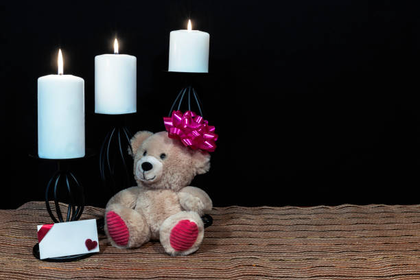 istock Cudlely teddy bear with pink bow on head, white candles perched on black candle holders on mesh place mat and wooden table with card and dark background. 1125191232