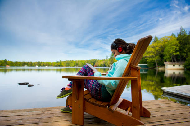 Teenager girl on a chair by the lake reading a tablet and enjoying music Teenager girl on a Muskoka chair reading a tablet and enjoying music on headphones. The girl is wearing colourful clothes. Lake cottages nestled between trees are visible in the background. cottage life stock pictures, royalty-free photos & images
