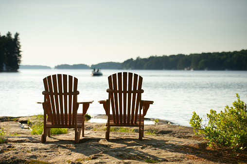 Adirondack chairs sit on a rock formation facing the waters of a lake during a sunny summer day in Muskoka, Ontario Canada. In the background a water boat is visible.