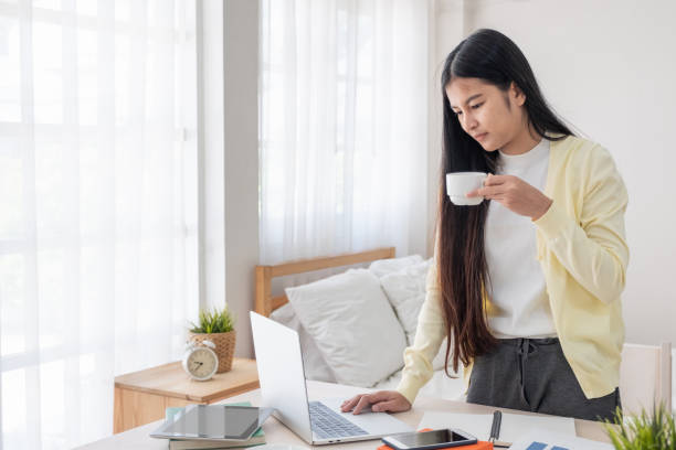 istock Asian female freelancer standing looking at laptop and drink coffee while working on table in bedroom at home.Work at home concept.work from home concept. 1125222117