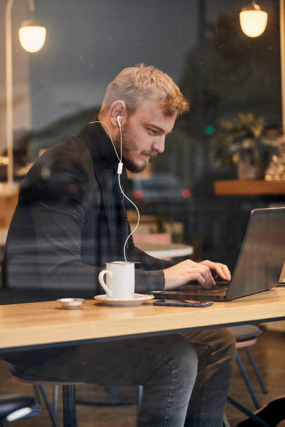 istock one young man, sitting in coffee shop and using his laptop while typing on keyboard. Shoot thought window outside (with reflections). 1125227172