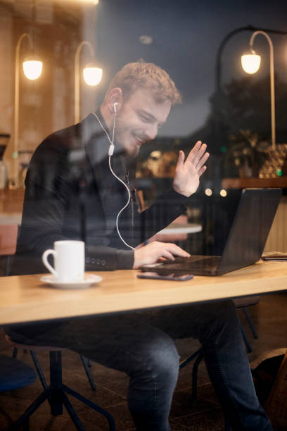 istock one young smiling man, sitting in coffee shop and using his laptop, gesturing with his hand Hello or Hi, waving his hand over video conference call. Shoot thought window outside (with reflections). 1125227241