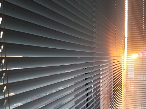 Closed window blinds with dawn sun bursting through, casting glow across slats