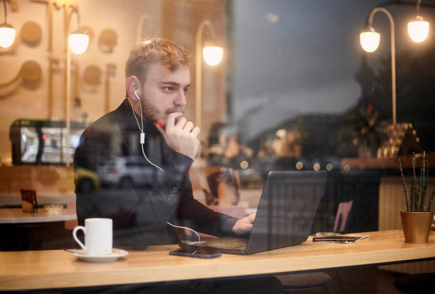 istock one young man, sitting in coffee shop and using his laptop while thinking and looking at street outside. Shoot thought window outside (with reflections). 1125231905