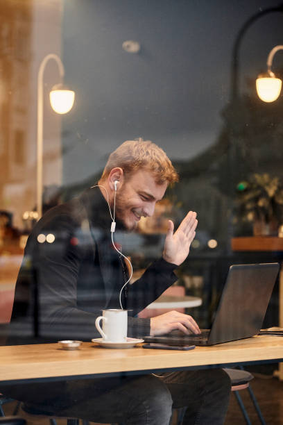 istock one young smiling man, sitting in coffee shop and using his laptop, gesturing with his hand Hello or Hi, waving his hand over video conference call. Shoot thought window outside (with reflections). 1125234547