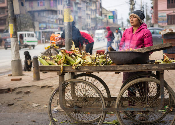 Kathmandu, Nepal - Jan. 27, 2019: A woman warms herself over her charcoal fire as she roasts corn for customers on the street. Nepal's economy is growing rapidly, but unequally, as the country struggles with a new constitution and growing industry. stock photo