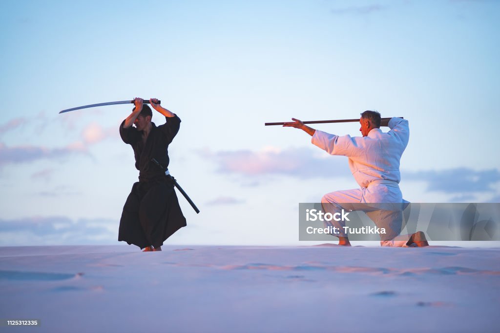Concentrated men, in Japanese clothes, are practicing martial arts Concentrated men, in Japanese clothes, are practicing martial arts with a traditional Japanese weapon - a katana and jo,  in the desert at sunset. Adult Stock Photo