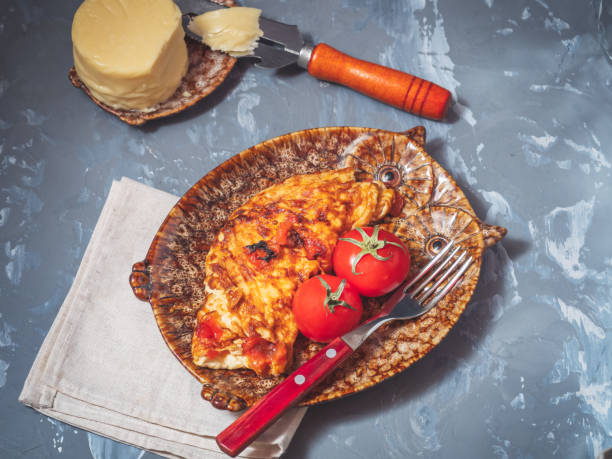 Breakfast mz omelet with vegetables and cheese. Decorative plates, two whole red tomatoes with leaves. A circle of cheese and a cheese knife in the background.