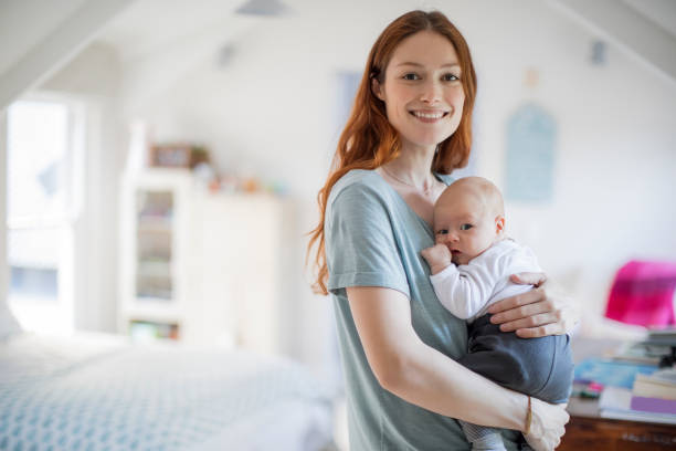 Smiling redhead mother carrying son at home Smiling redhead woman standing with cute son. Portrait of beautiful mother is carrying newborn baby boy in bedroom. They are at home. mom and baby eye contact stock pictures, royalty-free photos & images