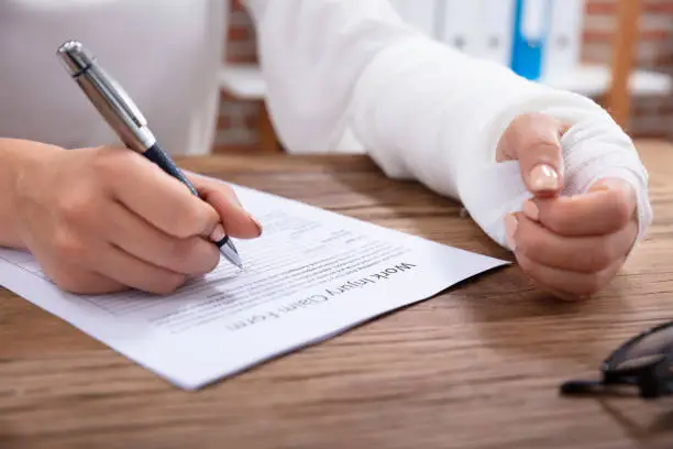 Close-up Of Businesswoman With White Bandage Hand Filling Work Injury Claim Form On Wooden Desk