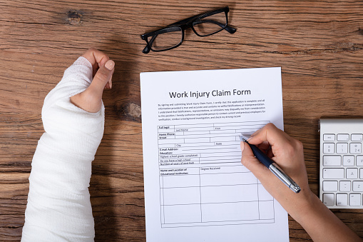 Close-up Of Businesswoman With White Bandage Hand Filling Health Insurance Claim Form On Wooden Desk