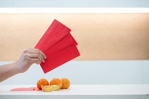 close up asian man hand holding red envelope over white table with mandarin oranges and gold ingots for special chinese new year traditional and culture concept
