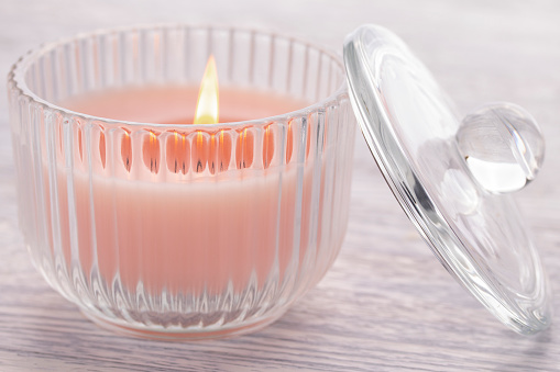 A pink candle is burning in a glass cup on an old white wooden table. Nearby is the lid of the glass