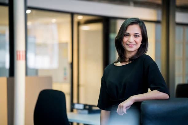 Smiling young businesswoman standing by chair Confident businesswoman standing by office chair at creative workplace. Portrait of smiling young female professional is in black casual. She is at office. smart casual stock pictures, royalty-free photos & images