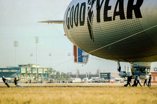 Pompano, Florida, USA – October 20, 1984 – A ground image of the  Goodyear Blimp Enterprise N1A, which was stationed at Pompano Air Park in South Florida. The Blimp color scheme logo was changed in 1991. The horizontal image shows crew holding harness lines with the blimp almost on the ground.