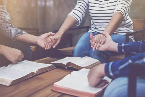 christian small group holding hands and praying together around wooden table with blurred open bible page in home room, devotional or prayer meeting concept