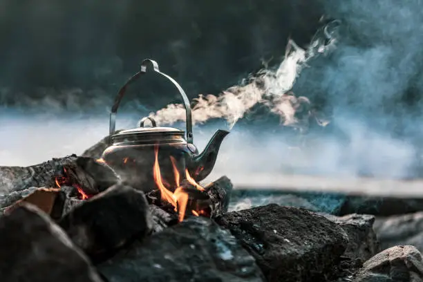 A close-up of a kettle boiling on a fire. There's smoke coming out from the kettle as well as from the fire.