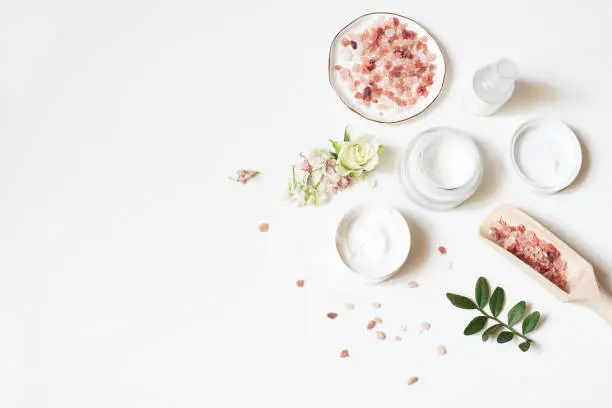 Styled beauty composition. Skin cream, tonicum bottle, dry flowers, rose and Himalayan salt on white table background. Organic cosmetics, spa concept, empty space, flat lay, top view.