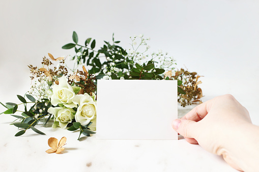 Bright feminine wedding stationery mockup scene. Closeup of woman hand holding paper place card. Bouquet of eucalyptus, dry hydrangea, white roses and gypsophila flowers. Marble table background.