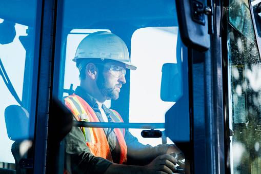 A mid adult man in his 30s working at a construction site, operating a backhoe. He is wearing a hardhat, safety vest and protective eyewear.