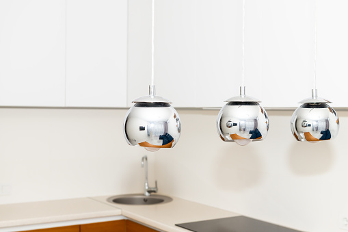 Closeup of three hanging ceiling lamps in kitchen with modern white cabinets in home house apartment or condo and reflection of living room in stainless steel luxury lights