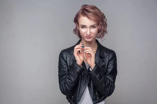 Portrait of cheater beautiful girl with short hairstyle, makeup in casual style black leather jacket standing with cunning idea and looking at camera. indoor studio shot, isolated on grey background.