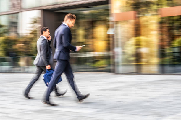 Blurred motion of two busy businessman walking in a hurry to glass office building, holding laptop bag and smart phone.