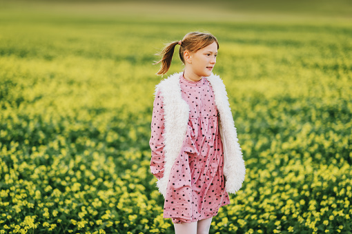 Outdoor portrait of pretty 10 year old girl, wearing pink vintage dress and faux fur bodywarmer, posing in a field