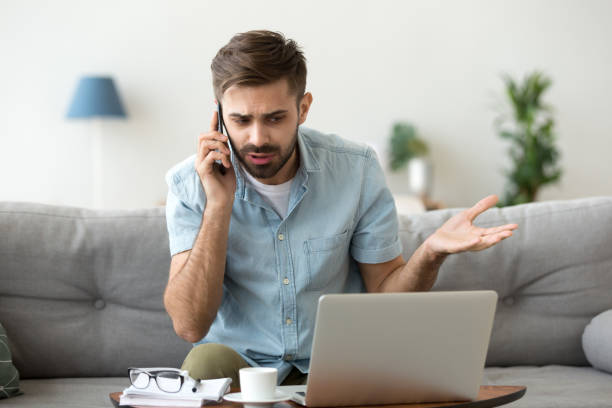 hombre enojado hablando por teléfono en disputa sobre el problema de la computadora portátil - quejándose fotografías e imágenes de stock