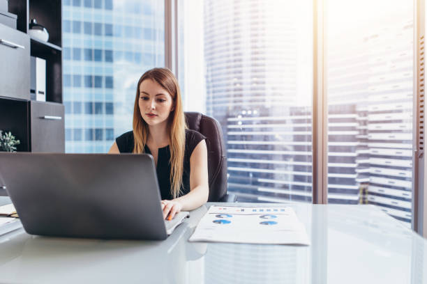 istock Female chief executive sitting at her desk taking notes in datebook writing with pen and using her computer in modern office building 1125578456