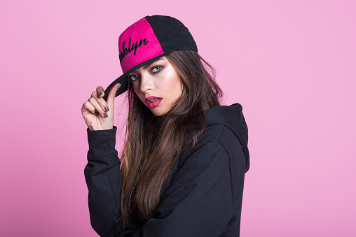 Studio portrait of beautiful young woman wearing black hooded shirt and baseball cap, standing against pink background and looking at camera.